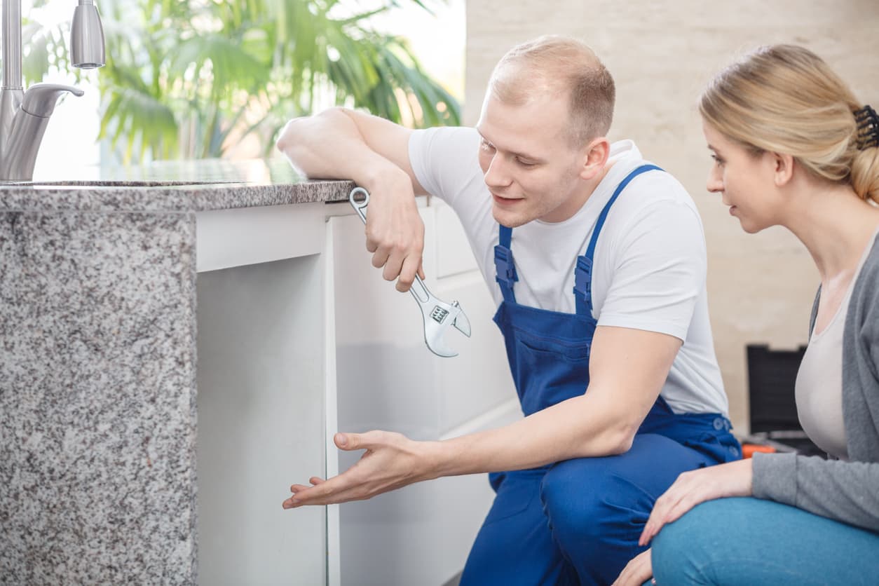 A plumber looking under a sink and talking to a woman