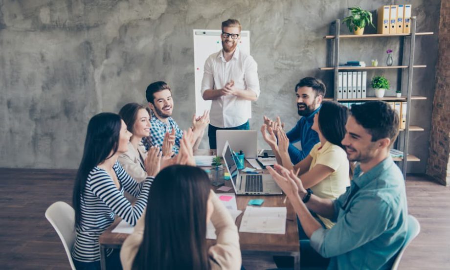 A group of people with laptops sitting around a table and applauding a man making a presentation.